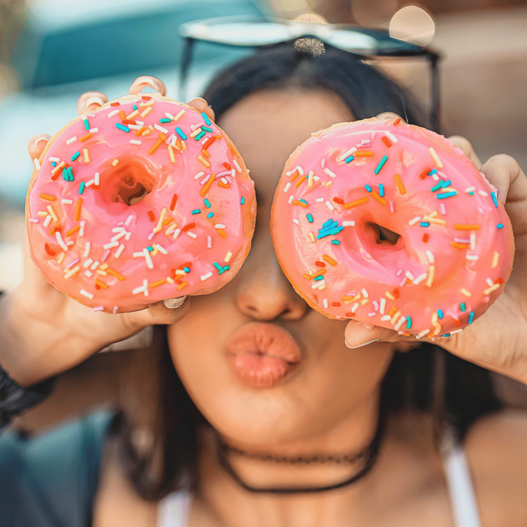lady holding two pink glazed donuts on her eyes and pouting